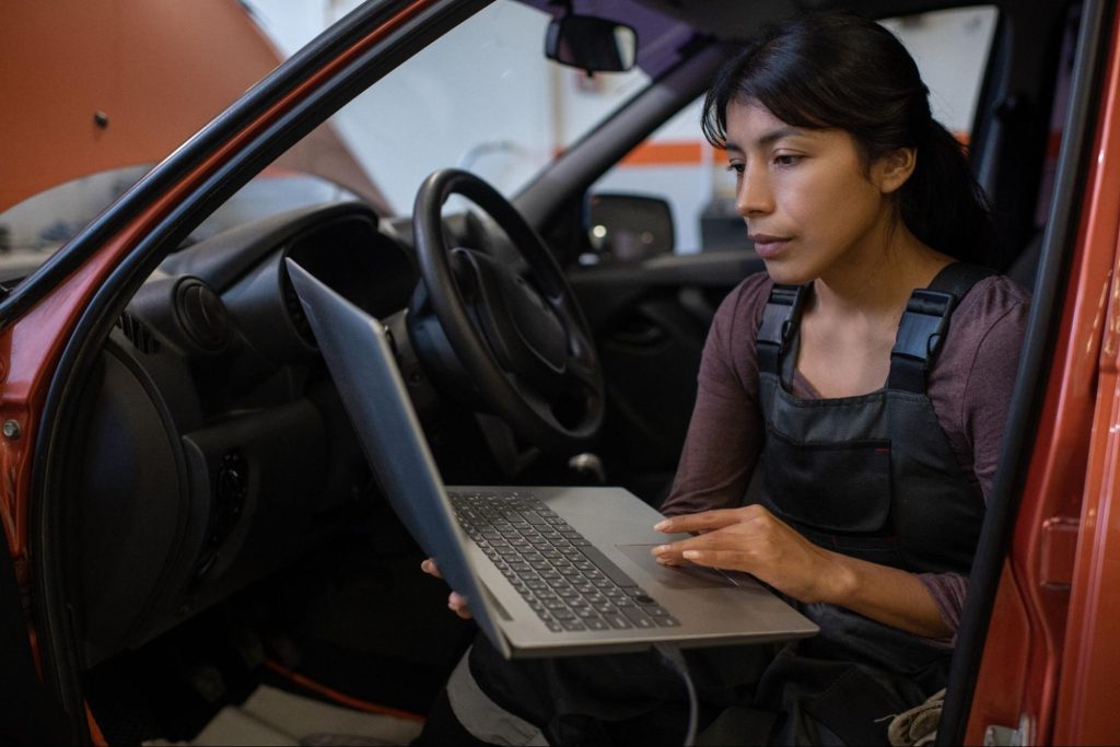 Auto technician sitting in driver seat runs diagnostics on a laptop during BCM repair service in a professional garage setting.