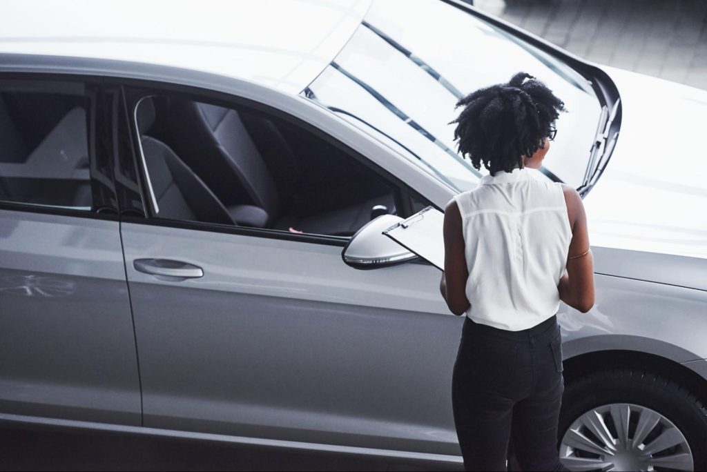 A female technician holding a clipboard examines a silver car in a dealership or garage setting, preparing to assess the vehicle’s car modules.